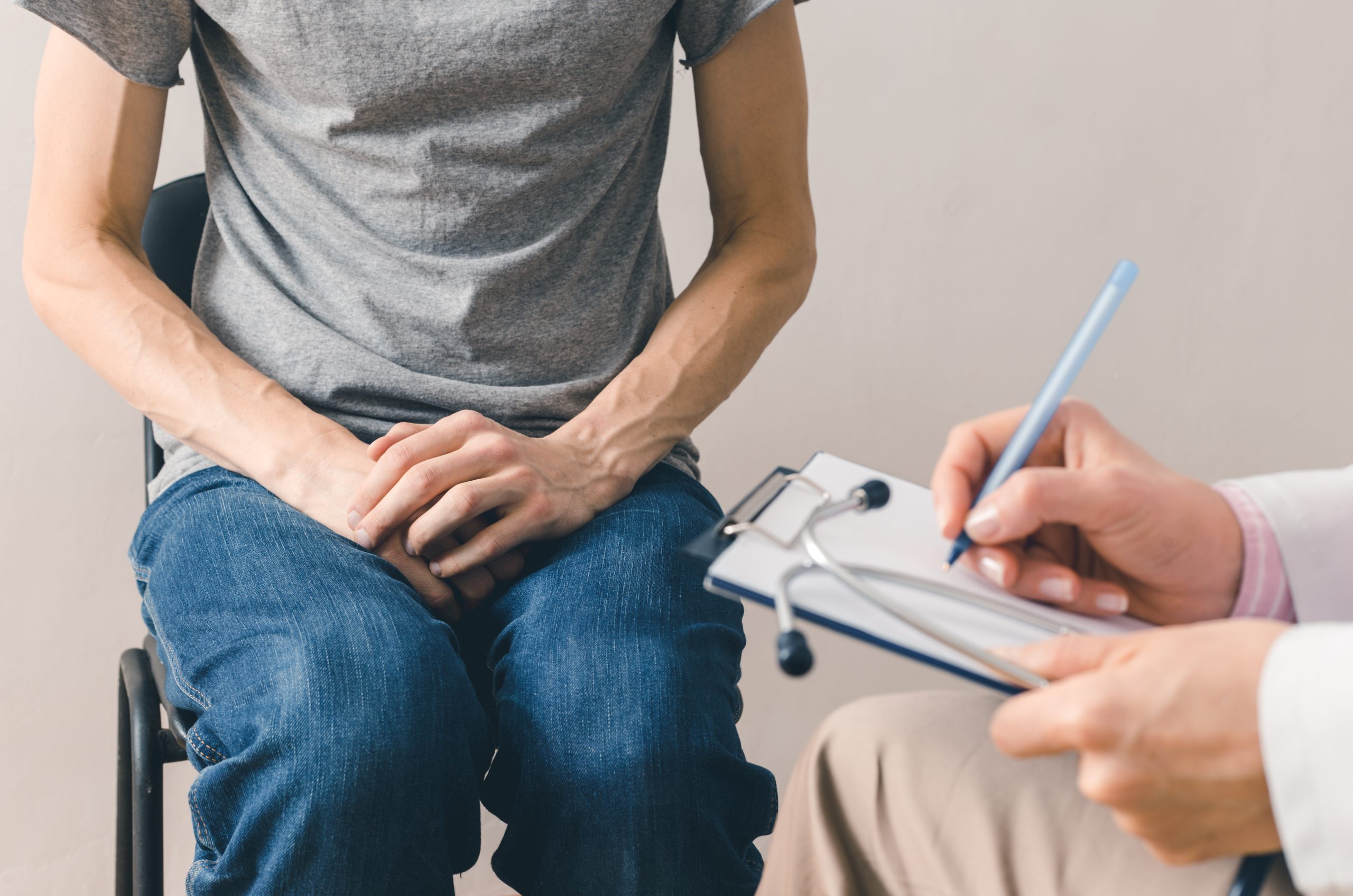 paciente sentado no consultório médico com a mão em cima do órgão genital masculino. Mãos aparente do médico segurando uma caneta e uma prancheta com papel